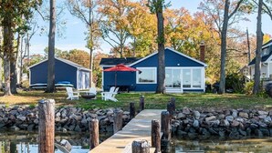 A lakeside cabin with a dock leading to a small boat, surrounded by trees and a stone wall. - Sunset Cottage - West River Access and Views! (Shady Side)