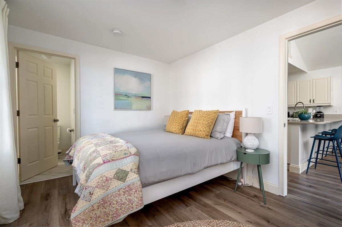 Een slaapkamer met een bed, een nachtkastje met een lamp, een schilderij aan de muur en uitzicht op een keuken.