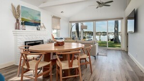 A spacious living room with a fireplace, a round wooden dining table with chairs, a sofa, and a large window offering a view of a lake. - Sunset Cottage - West River Access and Views! (Shady Side)