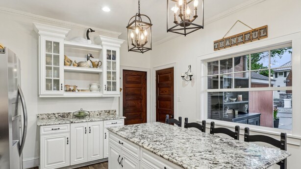 A kitchen with white cabinetry, a granite countertop, and a central island. There are two wooden doors and a window with a view of the outside. - Welcome to Grand Victorian on Main! (Ligonier)