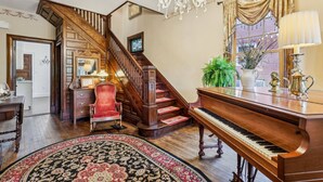 A grand room with a wooden staircase, a grand piano, and a chandelier. - Welcome to Grand Victorian on Main! (Ligonier)