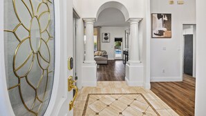Un corridoio con una porta in vetro decorativa, un pavimento piastrellato e vista su un soggiorno con divano e tavolo da pranzo.