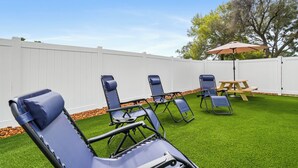 Un patio trasero con césped artificial, una valla blanca y varios sillones azules.