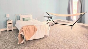 Un dormitorio con cama, mesita de noche, una pequeña mesa, una hamaca y una ventana con cortinas.