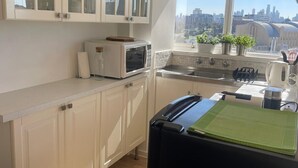 Une cuisine moderne avec des meubles blancs, un four à micro-ondes, une bouilloire et une vue sur la ville grâce à une grande fenêtre.
