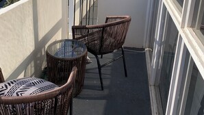 Un balcon agrémenté de meubles en osier et d’une table en verre, offrant une vue sur un paysage urbain avec ses bâtiments et sa verdure.