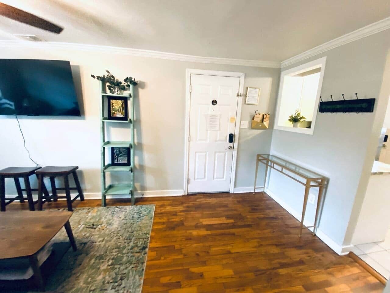 Une pièce avec un plancher en bois, une table à manger entourée de chaises, un téléviseur fixé au mur, une porte blanche et une petite étagère ornée d’objets décoratifs.