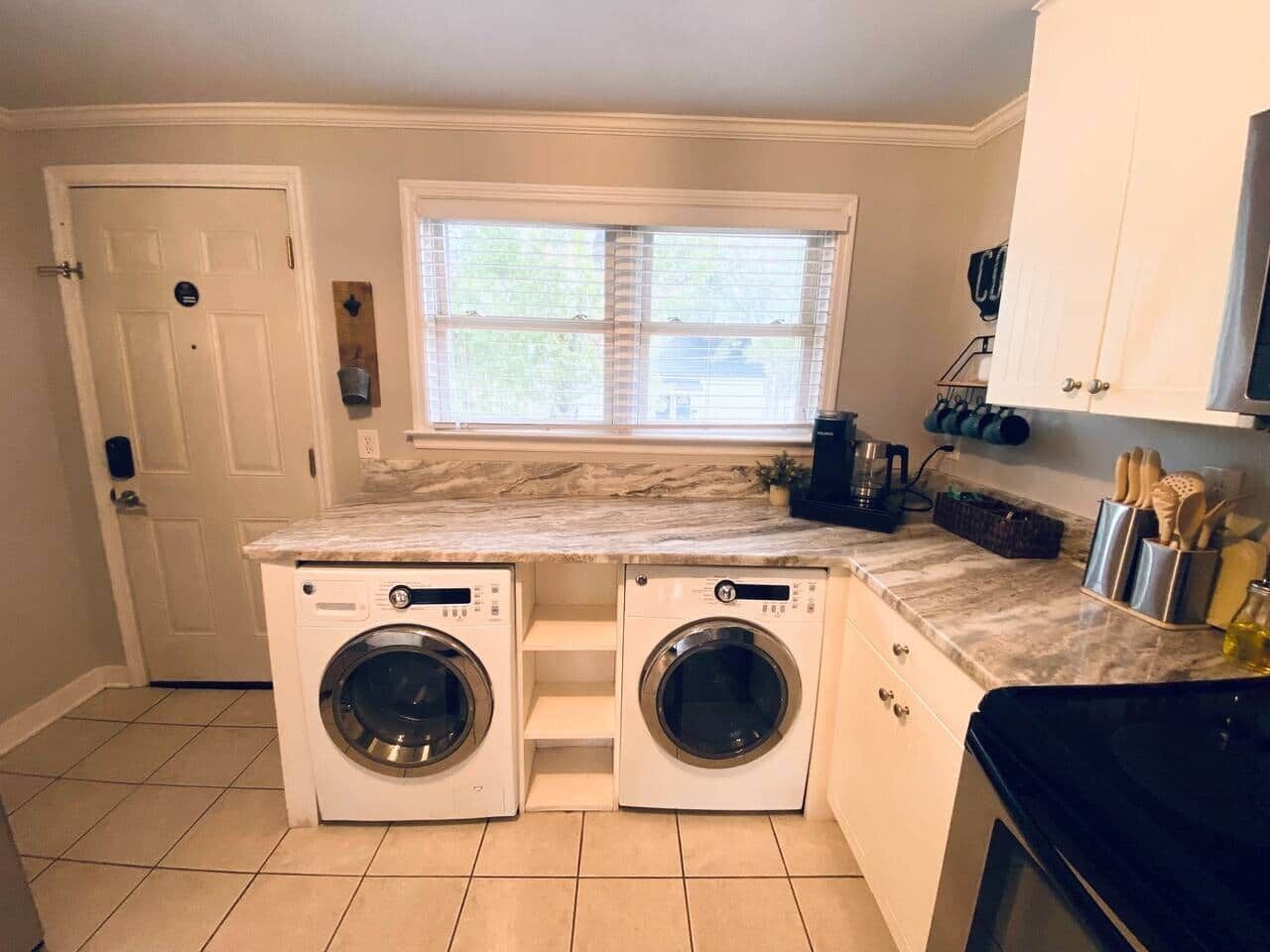 A laundry room with two front-loading washing machines, a window above the machines, and a countertop with various items.