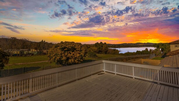 A wooden deck with a white railing, overlooking a lake and a grassy area with trees and houses in the background. - Lakeview Retreat in Elsinore with Pool & Hot Tub (Lake Elsinore)