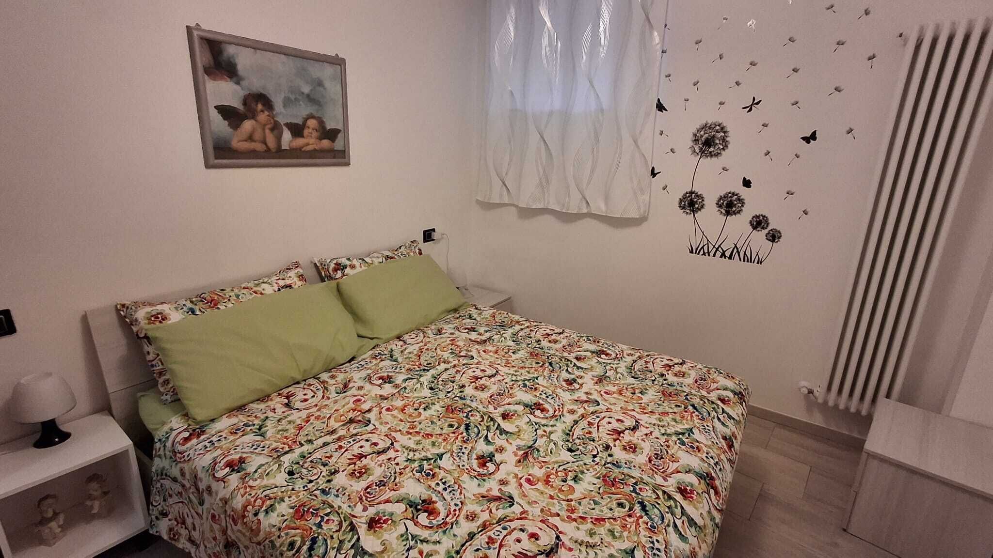 Een slaapkamer met een bloemenbedekking, een groen kussen, een nachtkastje met een lamp, een ingelijst schilderij aan de muur, en een muurdecoratie van bloemen en vlinders.