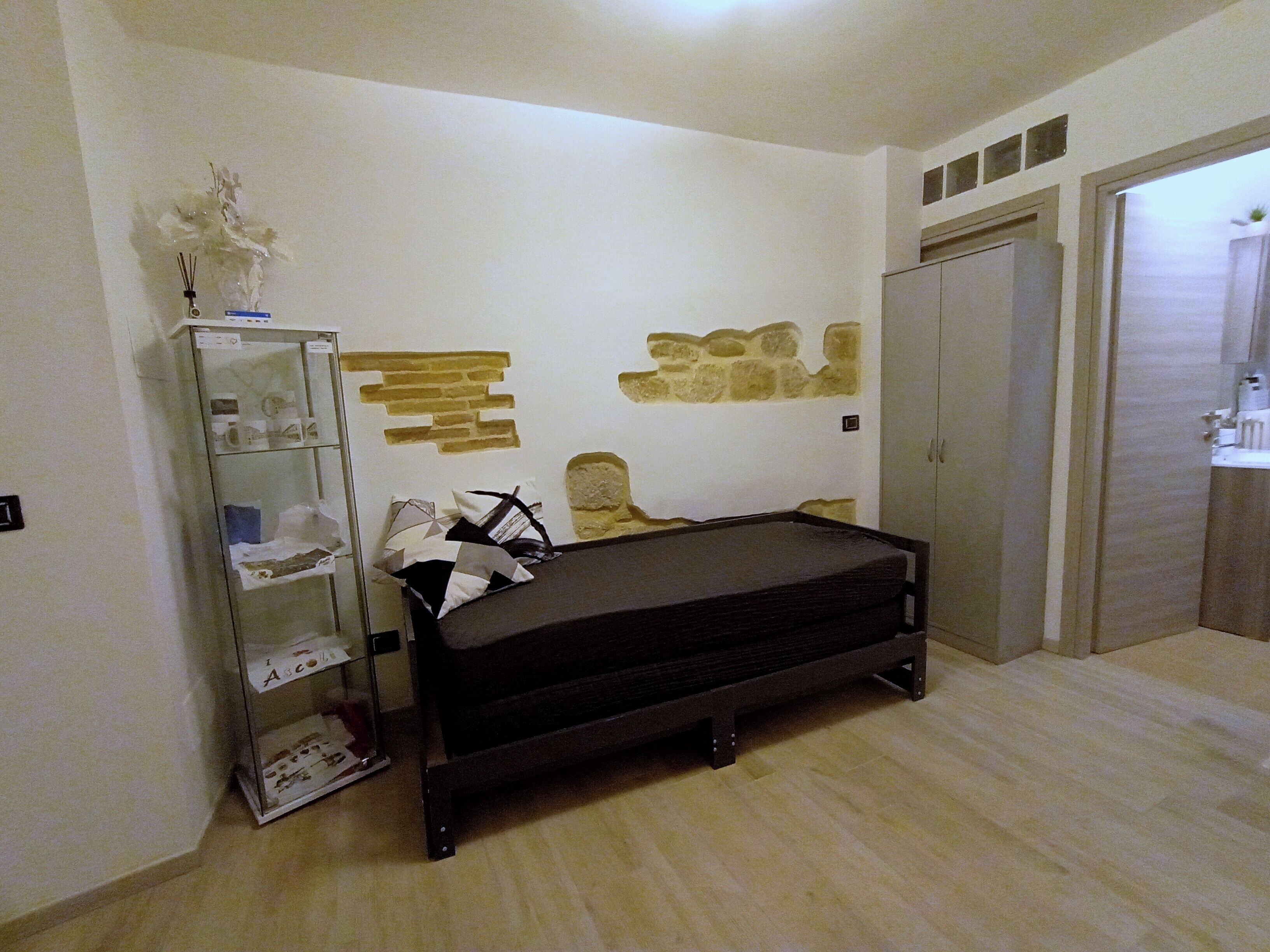 A bedroom with a bed, a wardrobe, a shelf with decorative items, and a wall with a stone-like design.
