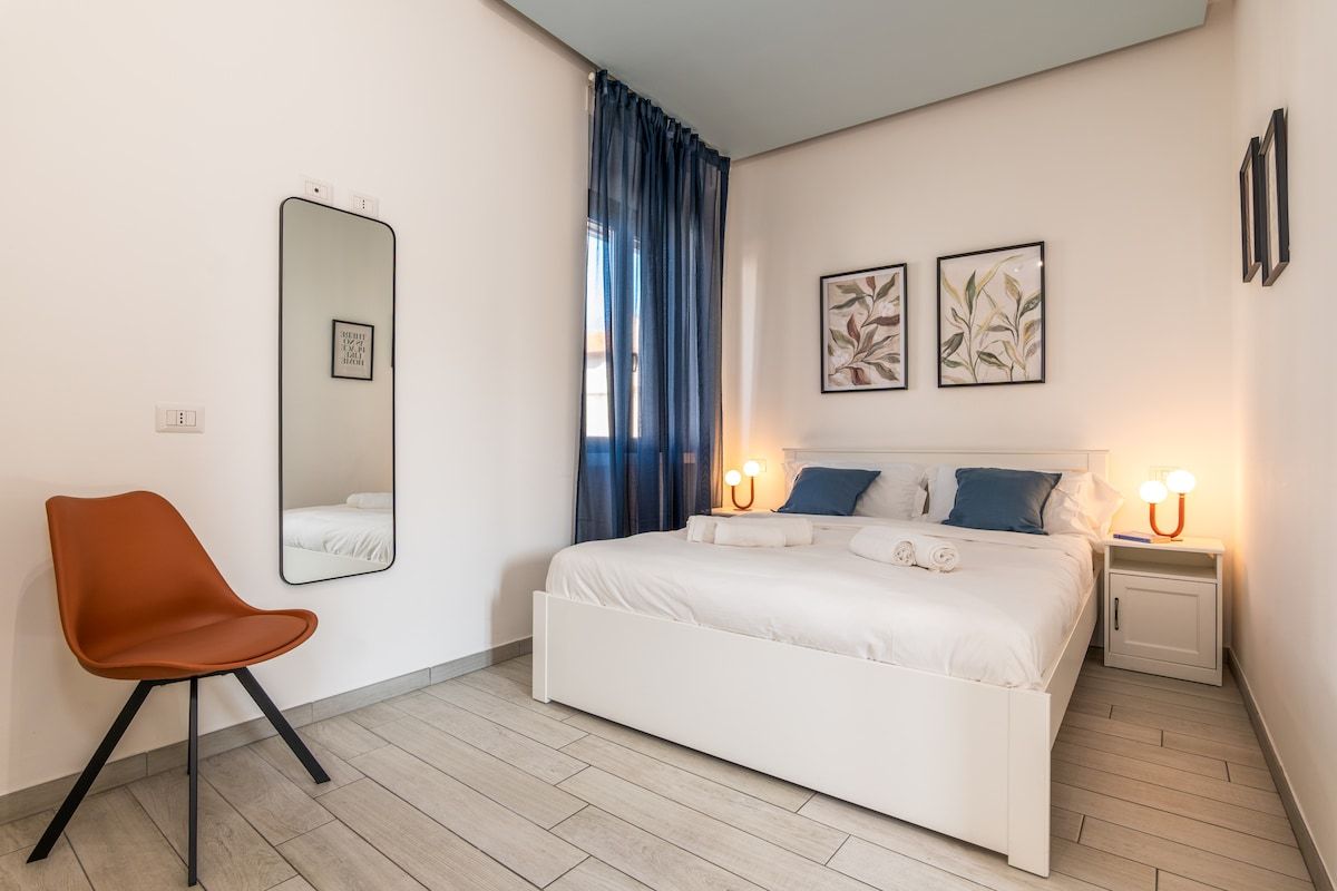 A bedroom with a bed, a chair, a bedside table, a mirror, and artwork on the wall.