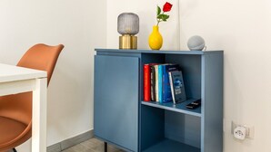 Ein modernes Zimmer mit einem flachbildfernseher an der Wand, einer blauen Kommode mit Büchern und Dekorationsobjekten, einem weißen Tisch mit einem braunen Stuhl und einem Holzboden.