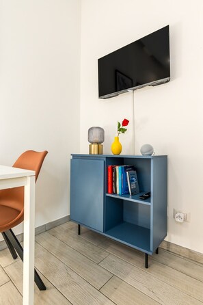 A modern room with a flat-screen TV mounted on the wall, a blue cabinet with books and decorative items, a white table with a brown chair, and a wooden floor. - Peaceful 1-Bedroom Flat | Steps from Metro (M1) (Milano)