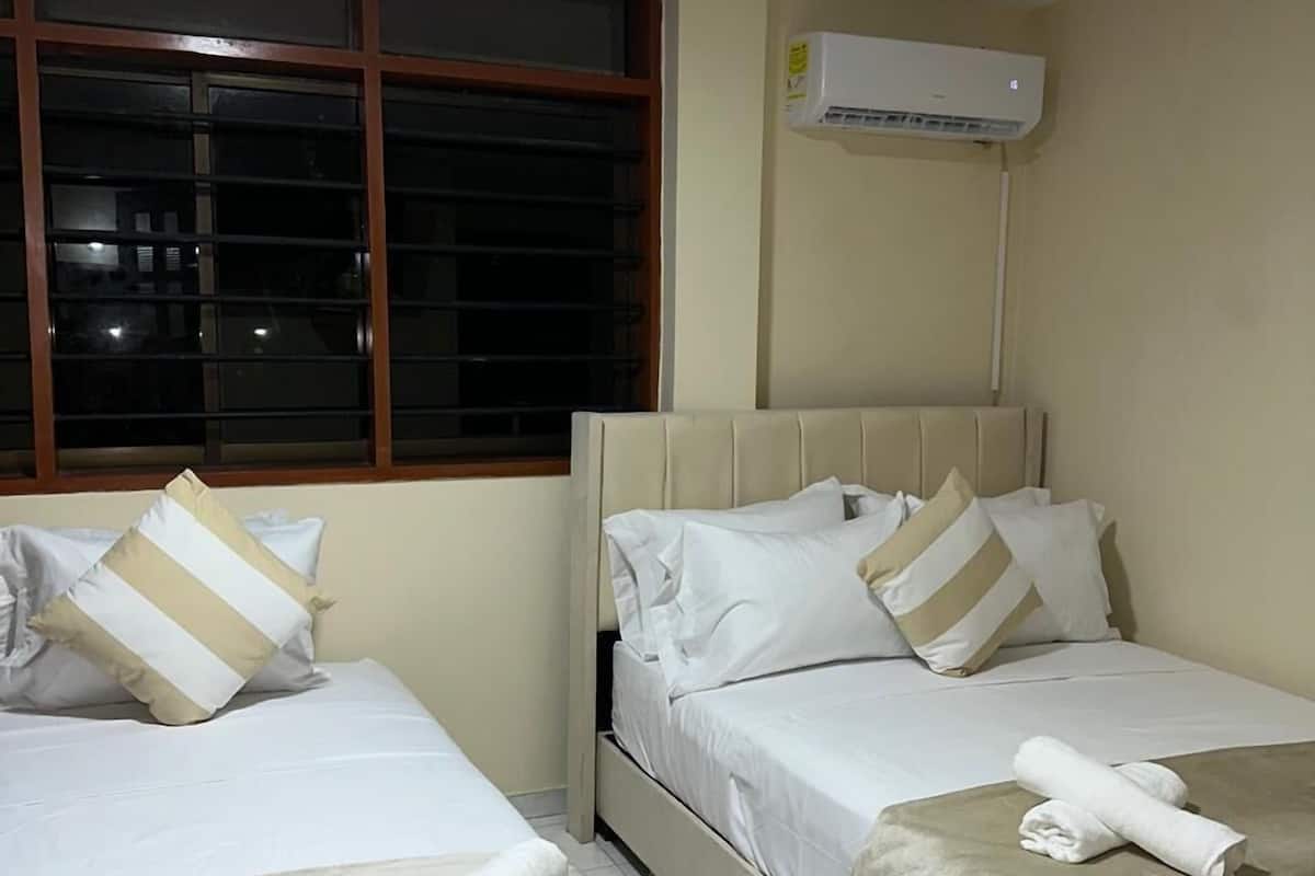 A hotel room with two beds, each with white bedding and striped pillows. There is a window with blinds, an air conditioning unit on the wall, and a tiled floor.