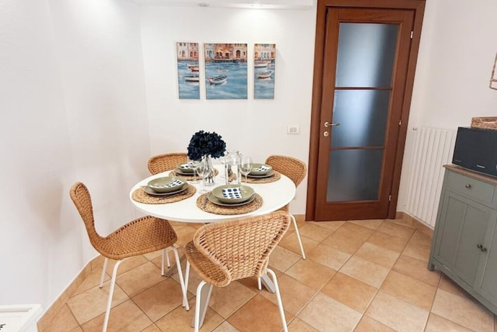 Uma sala de jantar com uma mesa redonda posta para quatro pessoas, rodeada por cadeiras de vime. Há três quadros na parede acima da mesa, retratando barcos na água. Uma porta com painéis de vidro jateado é visível à direita.