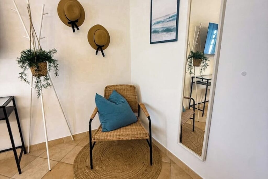 Une pièce comprenant un fauteuil en osier, un tapis rond tissé, un miroir et deux chapeaux accrochés au mur.
