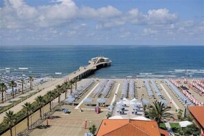Une plage bordée de rangées de parasols et de chaises longues, une jetée qui s’avance dans la mer et un ciel dégagé.