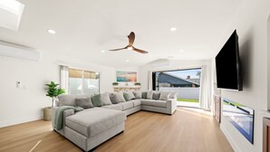 A modern living room with a sectional sofa, wooden flooring, a ceiling fan, and a large TV mounted on the wall. - IMPERIAL HOUSE (San Diego)