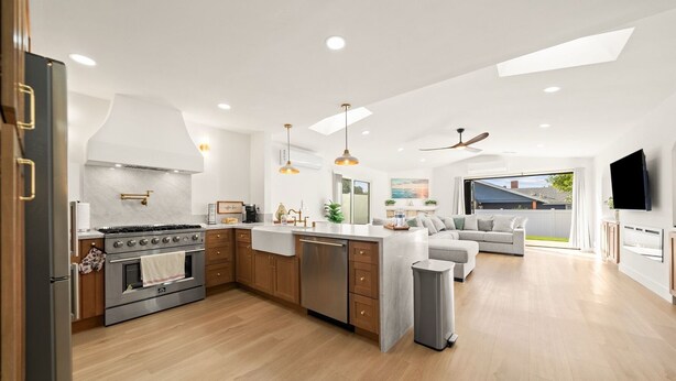 A modern kitchen with a central island, wooden cabinets, stainless steel appliances, pendant lighting, and a view to a backyard through large windows. - IMPERIAL HOUSE (San Diego)