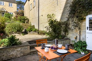 Un bâtiment en pierre avec une façade en briques, une porte blanche et un petit jardin agrémenté d’une table dressée pour un repas.
