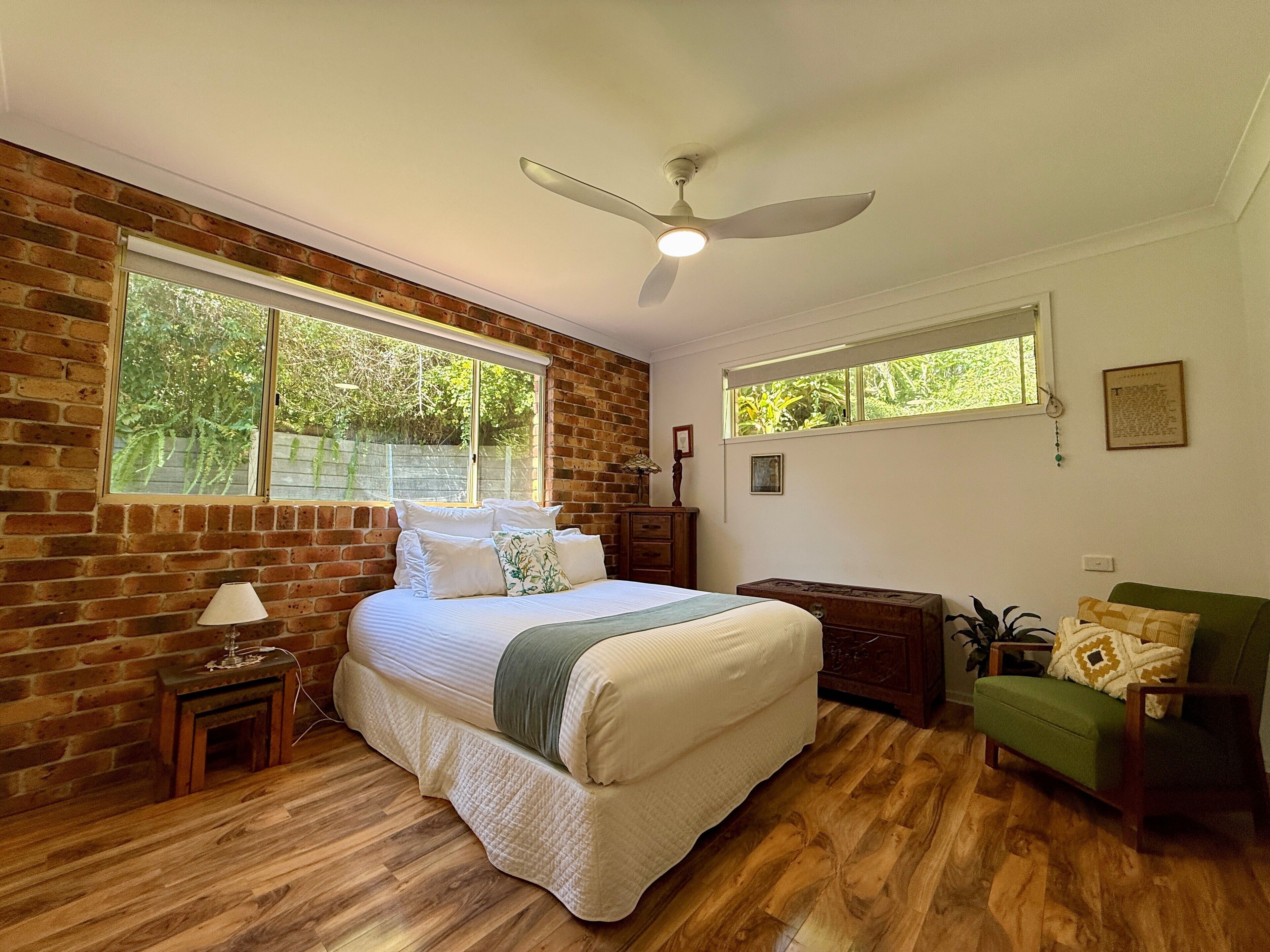 A bedroom with a brick wall, a large bed with pillows, a ceiling fan, a wooden dresser, a green armchair, and two windows providing natural light.