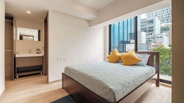 A modern bedroom with a large bed, a built-in wardrobe, a mirror, and a city view through large windows. - Brand New 2 BR Condo in Reforma - Queen Beds (Mexico City)