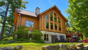 A log cabin with a stone foundation, large windows, and a deck with outdoor seating. - Chal’Heureux | Private Spa, Log Cabin Retreat at Lac Fiddler | Dog Friendly (Mille-Isles)