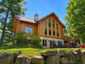 A log cabin with a stone foundation, large windows, and a deck with outdoor seating. - Chal’Heureux | Private Spa, Log Cabin Retreat at Lac Fiddler | Dog Friendly (Mille-Isles)