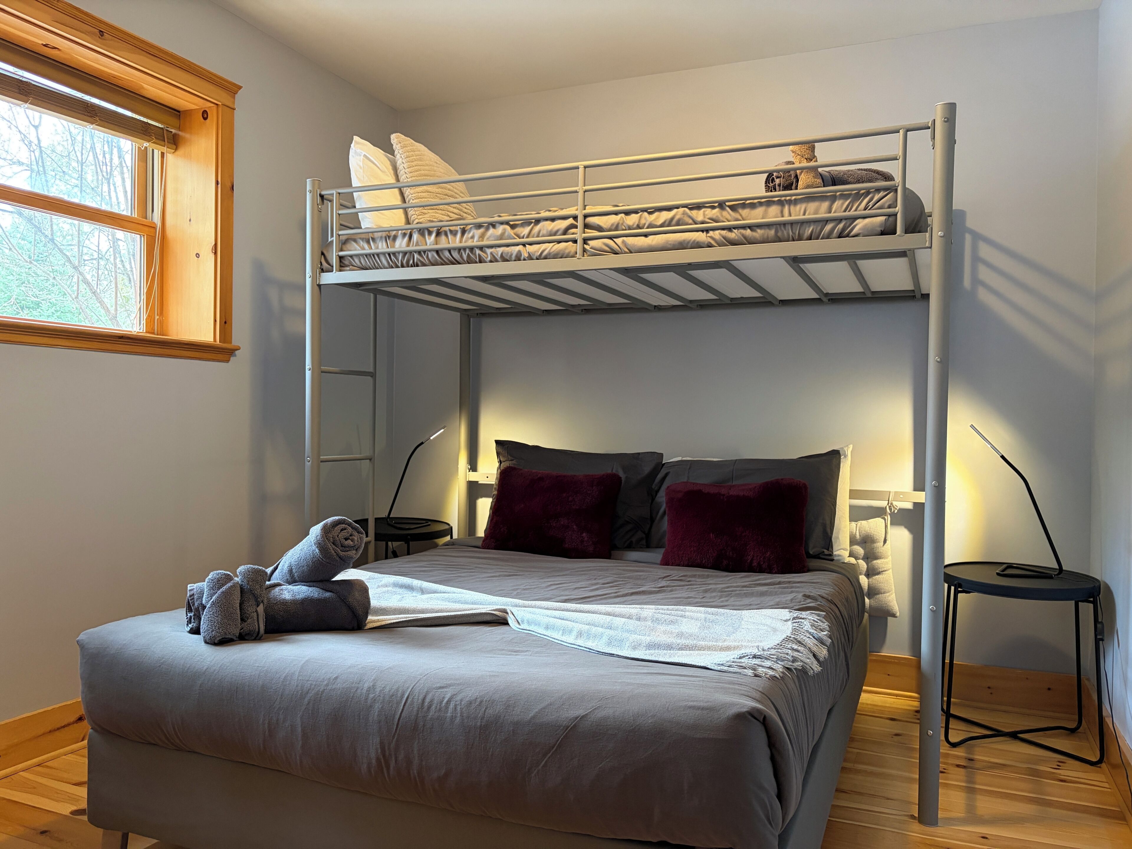 Un lit superposé avec un bureau et une chaise, une fenêtre avec des stores et une lampe sur une table de chevet.