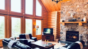 A log cabin interior with a stone fireplace, leather furniture, and a television. - Chal’Heureux | Private Spa, Log Cabin Retreat at Lac Fiddler | Dog Friendly (Mille-Isles)