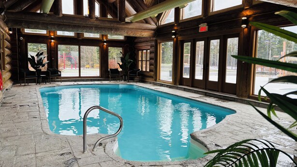 An indoor swimming pool with stone flooring, wooden beams, and large windows. - Chal’Heureux | Private Spa, Log Cabin Retreat at Lac Fiddler | Dog Friendly (Mille-Isles)