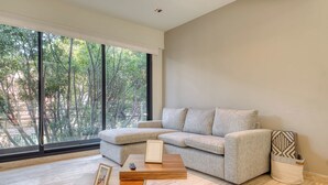 A modern living room with a sectional sofa, a wooden coffee table, and a large window offering a view of trees. - Centric 2 BR Condo in the Heart Roma Norte (Mexico City)