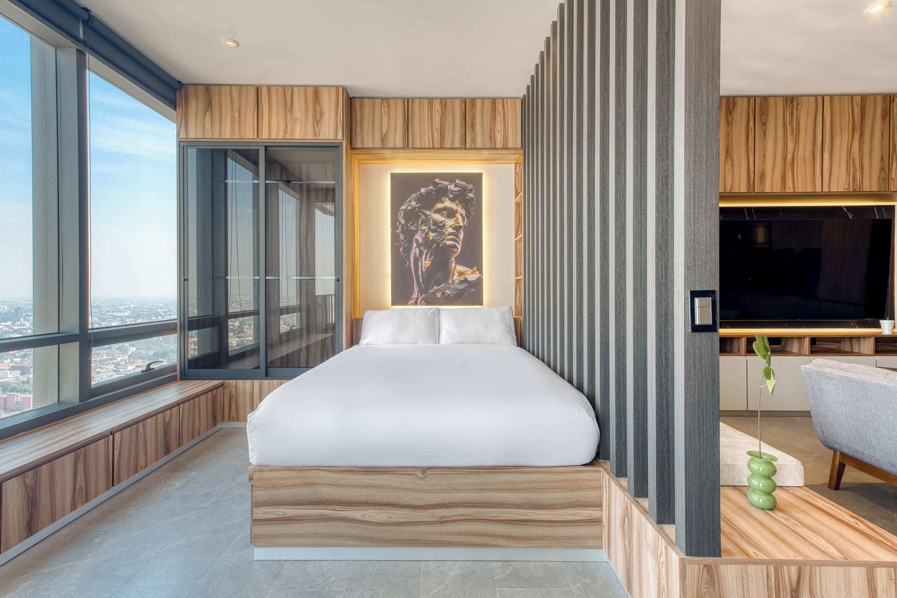 A modern bedroom with a large bed, a city view, a wall-mounted TV, and a green plant.