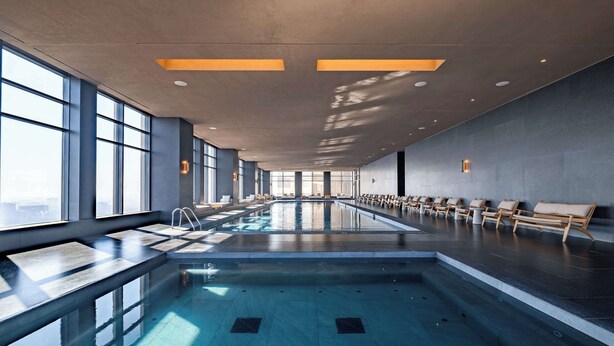 A modern indoor swimming pool with clear glass walls, a long bench seating area, and a view of the city skyline. - Luxury Studio - Pool - Revolution Monument View (Mexico City)