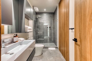 A modern bathroom with a large white sink, a toilet, a glass-enclosed shower, and wooden doors. - Luxury Studio - Pool - Revolution Monument View (Mexico City)