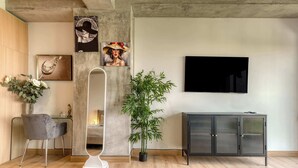 Ein modernes Wohnzimmer mit Holzboden, einem grauen Sessel, einer hohen Stehlampe, einem Blumentopf, einer schwarzen Kommode und einem flachbildfernseher an der Wand.