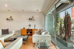A modern living room with a glass coffee table, white sofas, a wooden cabinet, and a large window with a view of cacti. - Centric 2 BR Condo - Roma - Rio de Janeiro Park (Mexico City)