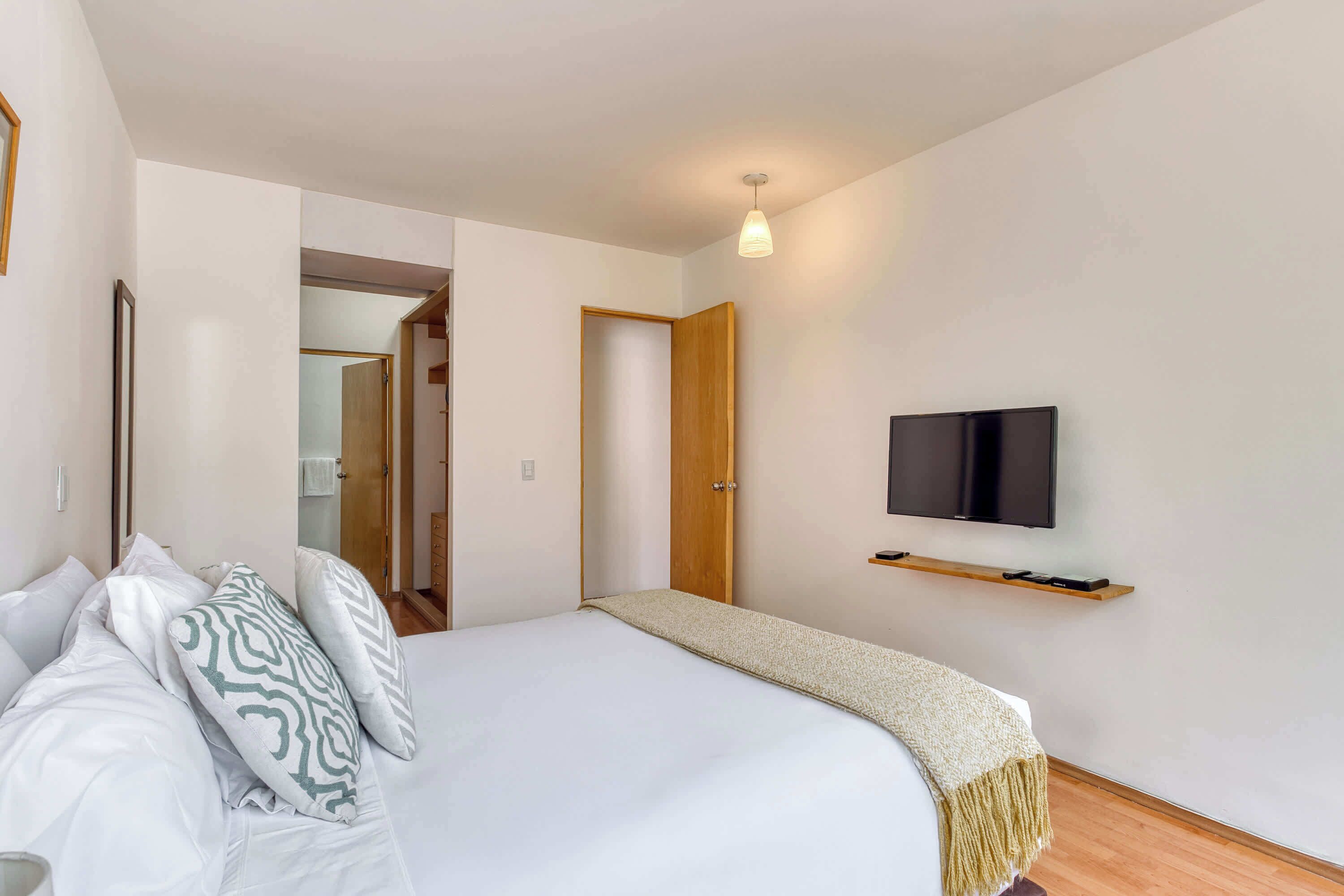 A bedroom with a bed, pillows, a TV, a wooden shelf, and a door leading to another room.