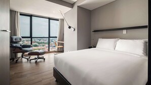 Et moderne hotelværelse med en stor seng, et skrivebord med kontorstol og udsigt over byen gennem store vinduer.