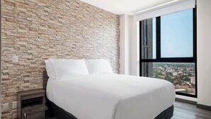 Une chambre à coucher moderne avec un mur au motif de pierres, un grand lit avec du linge de lit blanc, une table de chevet et une vue sur le paysage urbain par une grande fenêtre.
