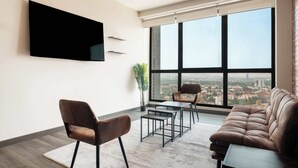 Un salon moderne doté d’une grande fenêtre, d’un téléviseur à écran plat, d’un canapé, de deux fauteuils et d’une petite table.
