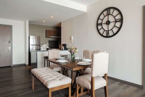 Un comedor moderno con una mesa de madera, sillas acolchadas, un reloj de pared grande y una cocina al fondo.