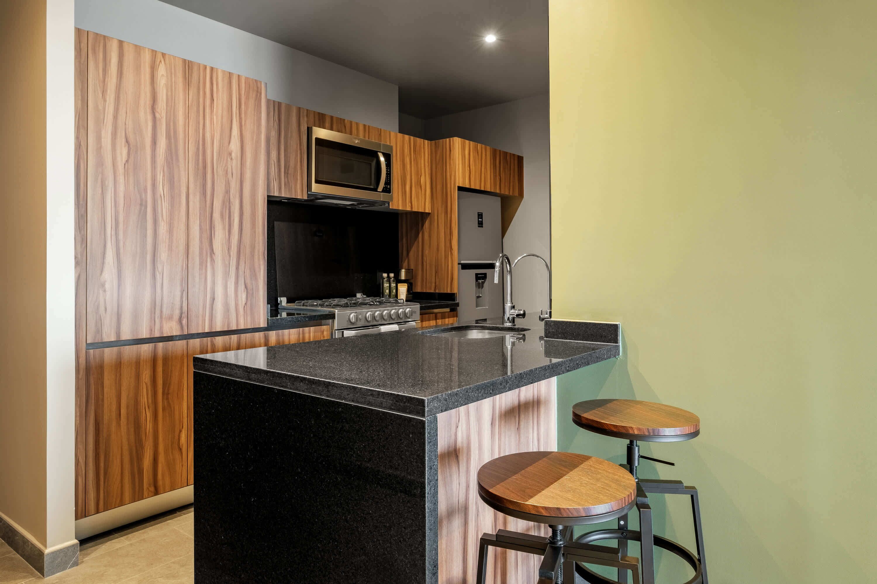 A modern kitchen with a central island, wooden cabinetry, a microwave, a stove, and two stools.