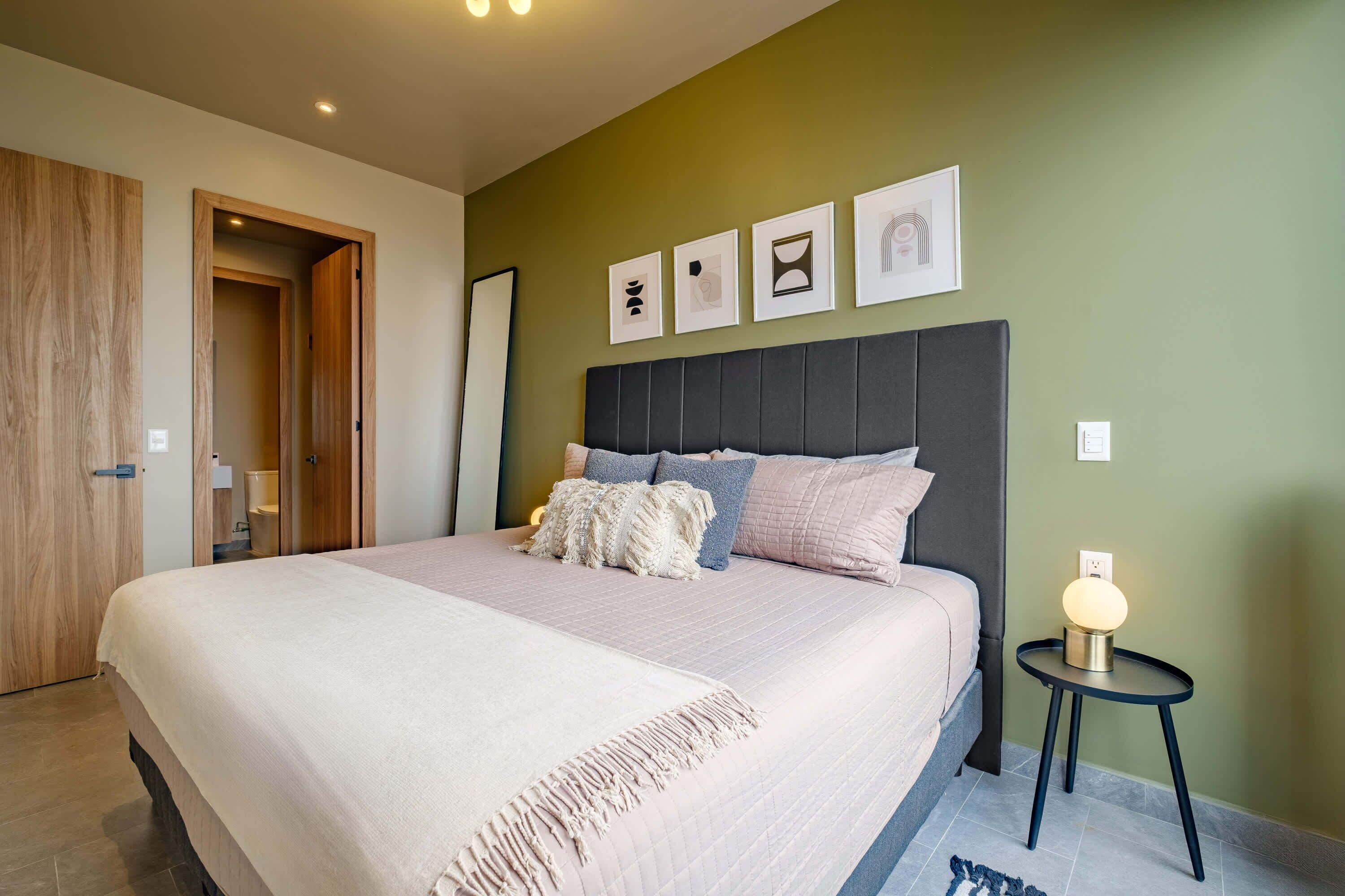 A bedroom with a large bed, a bedside table with a lamp, a door, and a wall with framed pictures.