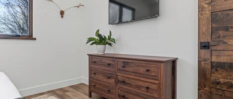 A bedroom with a wooden dresser, a flat-screen TV mounted on the wall, a window with a view of bare trees, and a door with a wooden panel.