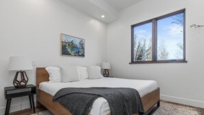 Une chambre avec un lit en bois, une literie blanche, un plaid gris, un tableau au mur et une fenêtre donnant sur des arbres.