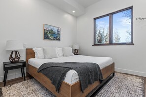 Ein Schlafzimmer mit einem hölzernen Bett, weißer Bettwäsche, einer grauen Tagesdecke, einem Gemälde an der Wand und einem Fenster mit Blick auf Bäume.