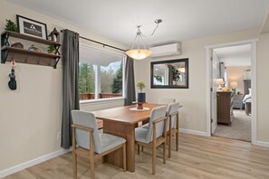 A dining area with a wooden table and chairs, a hanging light fixture, and a view through a window. - Hood Canal  Retreat: Hot Tub, Water Views, Fire Table Fully Stocked Kitchen (Hoodsport)
