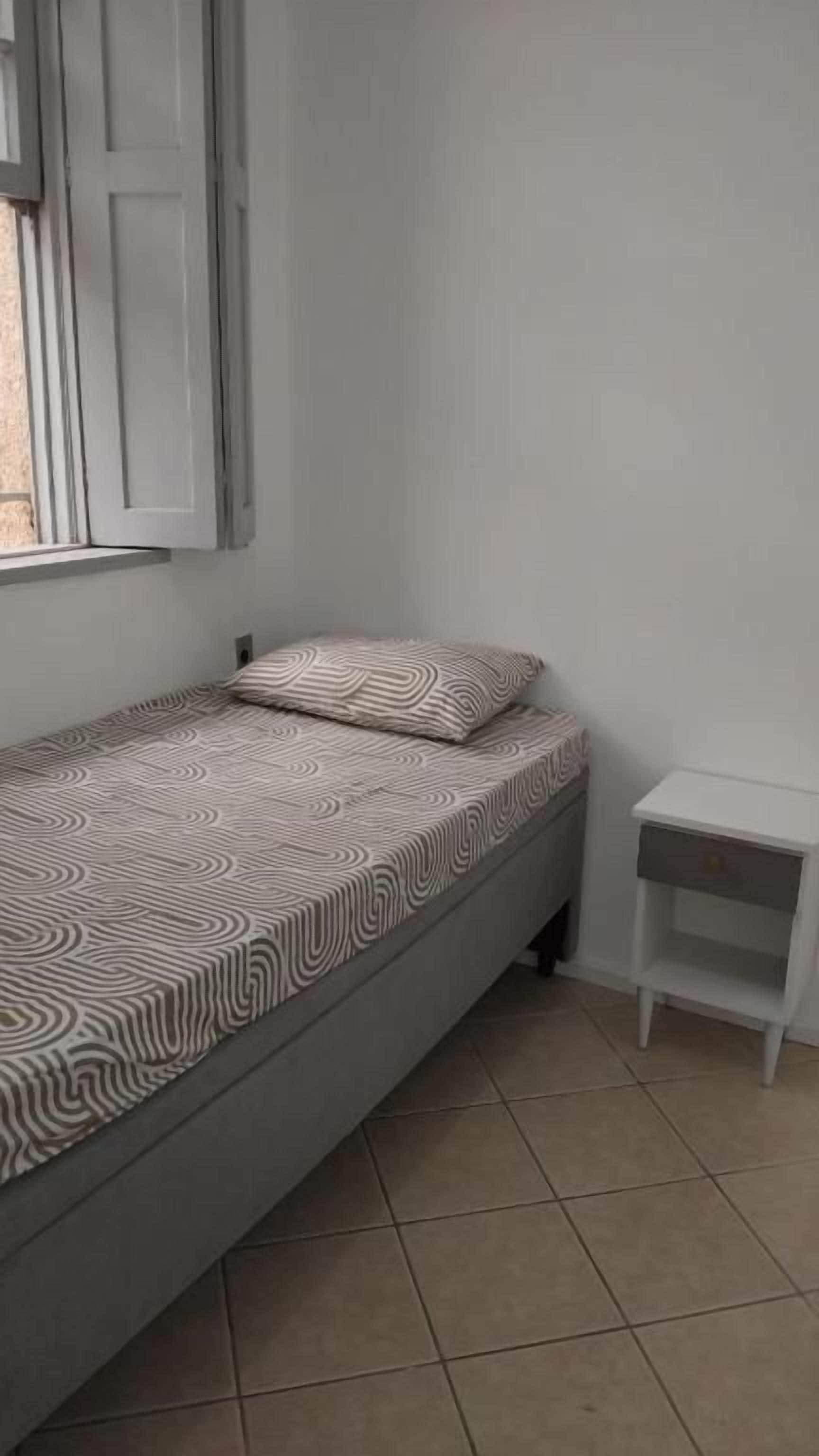 A single bed with patterned bedding, a small bedside table, and a window with shutters.